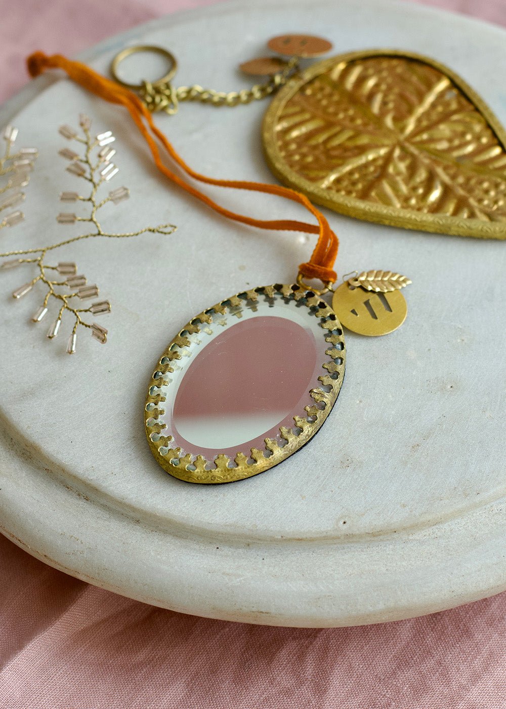 Oval ex voto mirror ornament with golden frame and orange velvet ribbon, styled on a marble plate with pink fabric background.