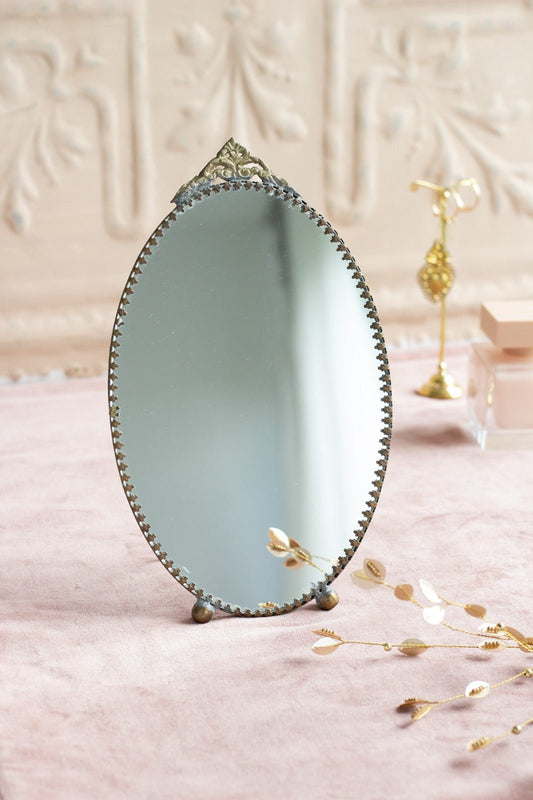 Oval vintage-style tabletop mirror with decorative brass detailing and ornate top, displayed on a soft pink surface with golden accessories.