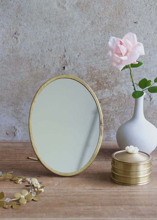Oval tabletop mirror with a brass frame and minimal stand, styled with a white vase, a pink rose, and golden accessories on a wooden surface.