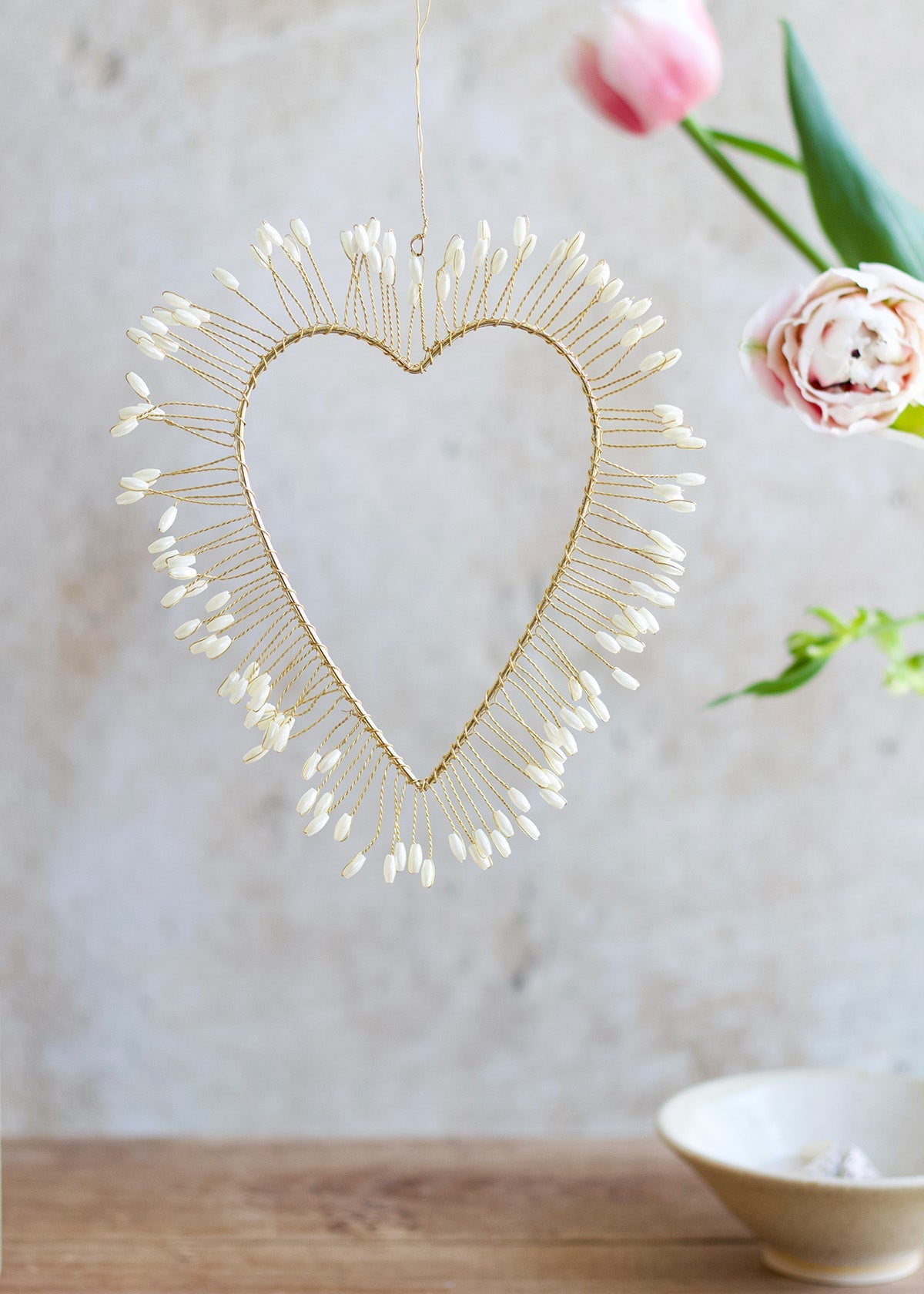 Delicate heart-shaped ornament made of gold wire and ivory-colored buds, hanging in front of a soft background with tulips.