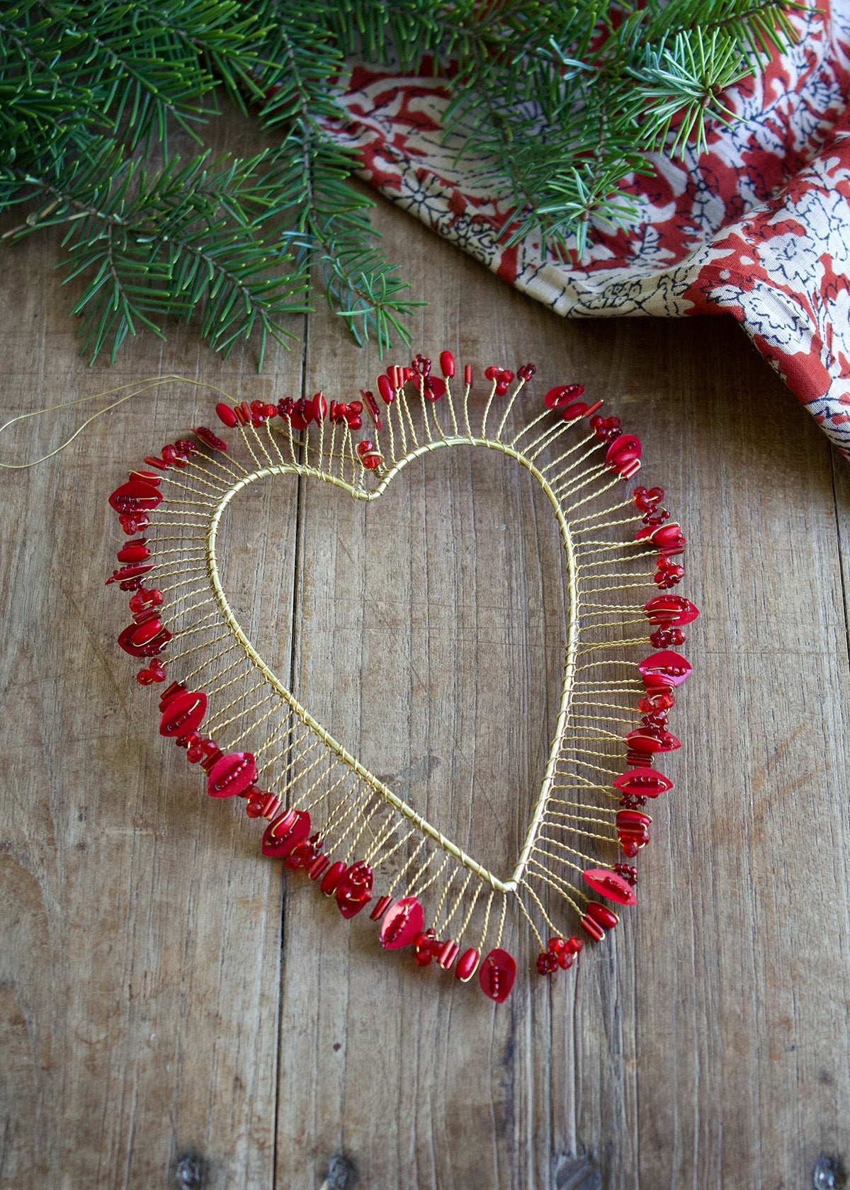 Heart-shaped decorative piece with red beads on a wooden surface with greenery and fabric in the background