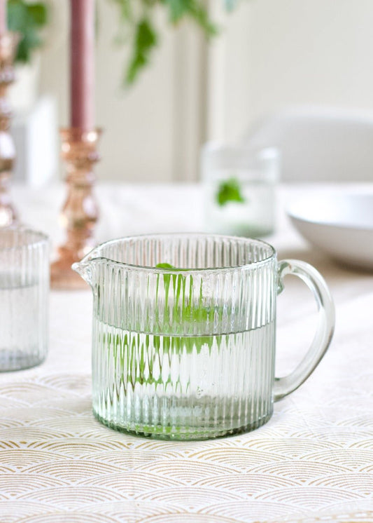 Low clear glass jug with vertical ribbed texture and round handle, placed on a softly patterned tablecloth.