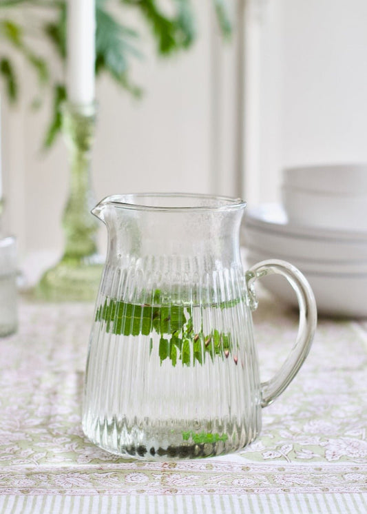 Clear ribbed glass pitcher filled with water and mint, placed on a patterned tablecloth with white bowls and green glassware in the background.