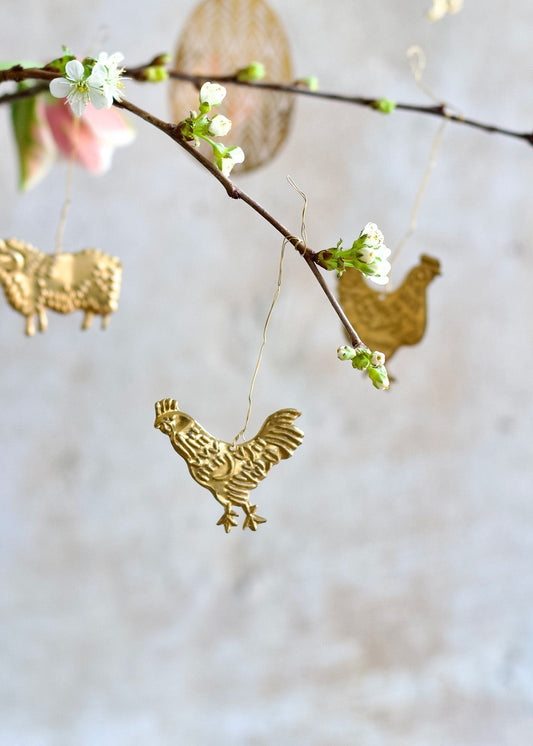 Brass rooster ornament hanging from a blossoming branch, with other golden animal shapes in the background.