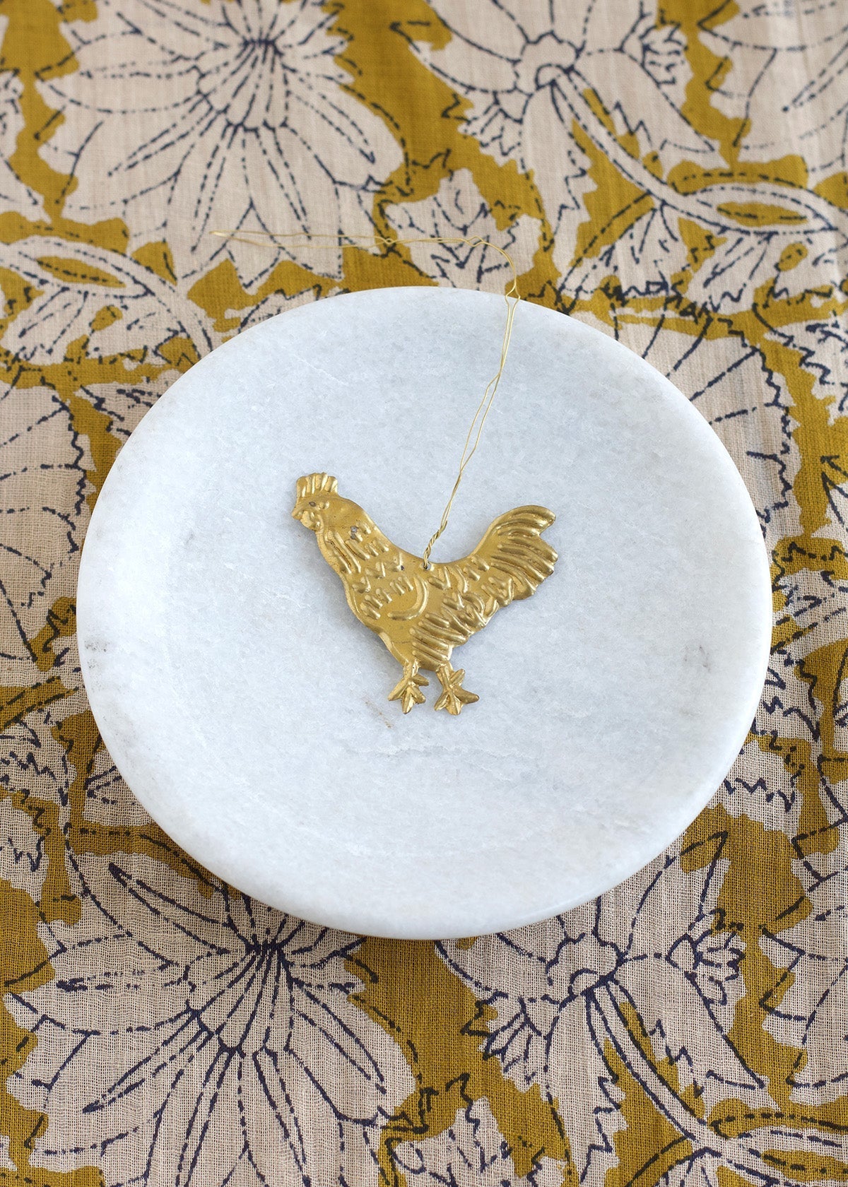 Brass rooster ornament displayed on a round white marble dish with floral mustard-yellow fabric in the background.