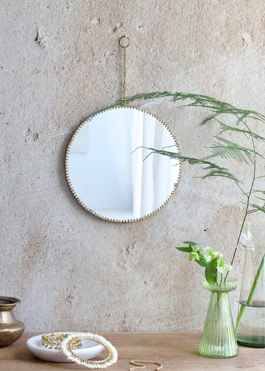 Round mirror with decorative frame hanging on a textured wall with plants and a vase below.