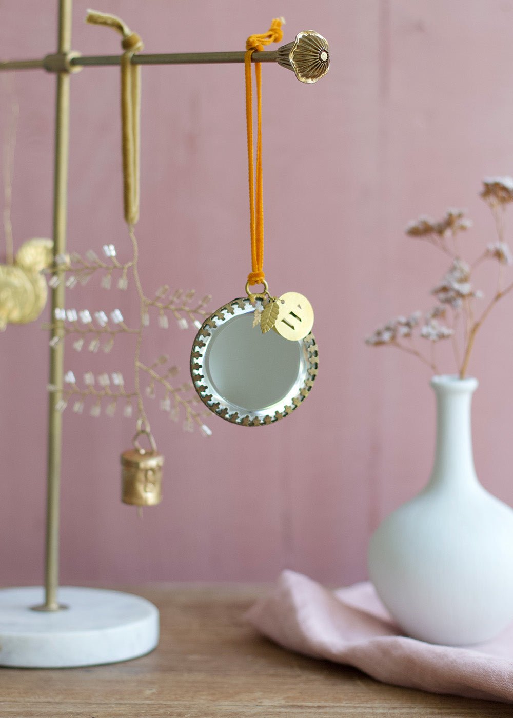 Round ex voto mirror ornament with golden frame and orange velvet ribbon, styled on a brass ornament stand with a pink background.