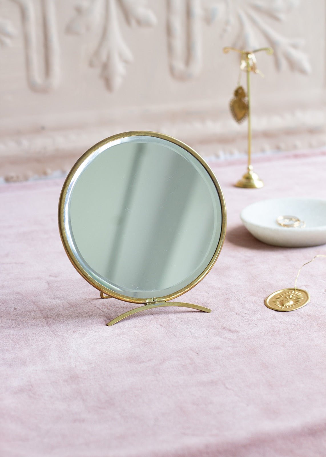 Round table mirror with brass frame and curved stand, placed on a soft pink fabric with golden jewellery and decor in the background.