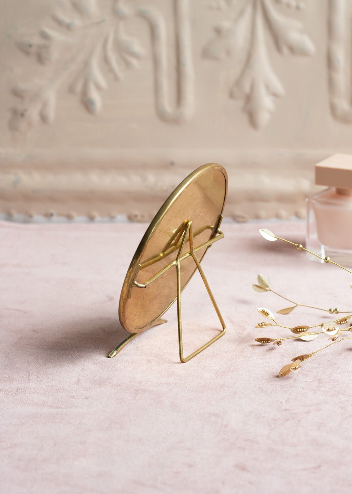 Back view of a round tabletop mirror with a brass frame and elegant wire stand, placed on soft pink fabric next to golden decor elements.