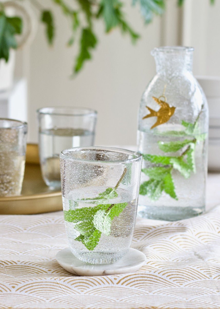 Clear glass with water and mint leaves, goldfish in a jar, on a patterned tablecloth.