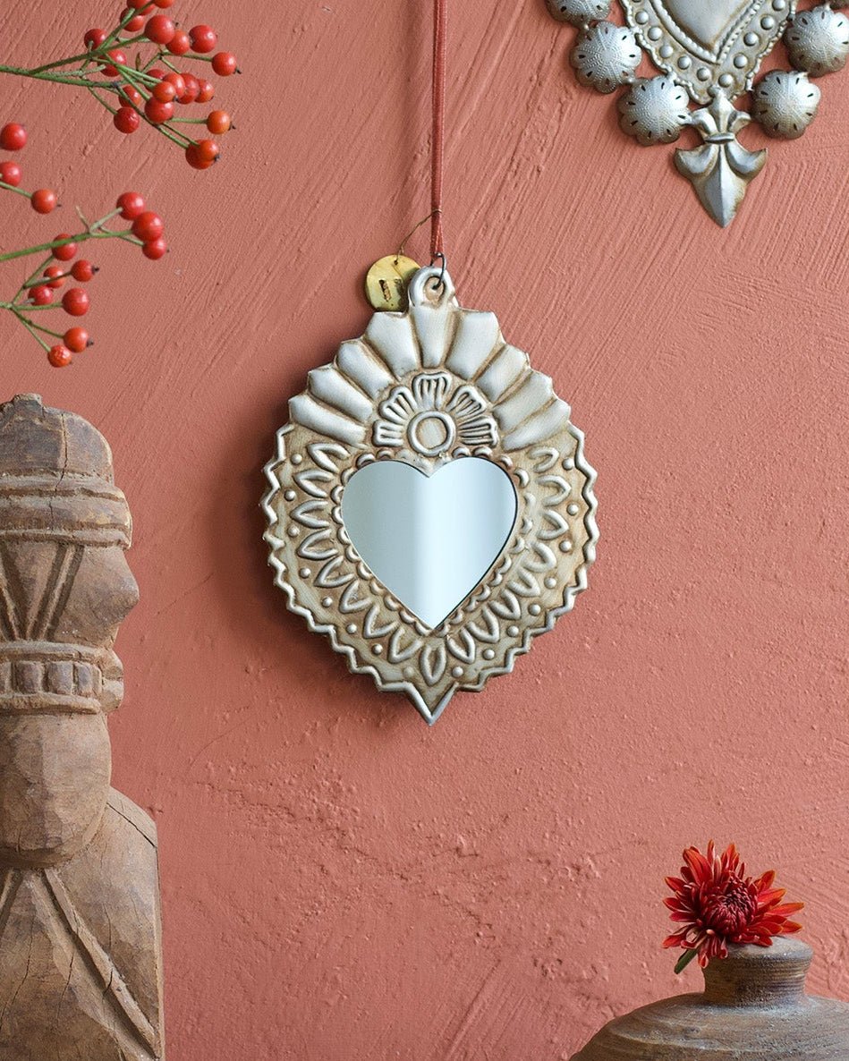 Silver heart-shaped mirror with embossed metal frame hanging on a terracotta wall beside wooden and ceramic objects.