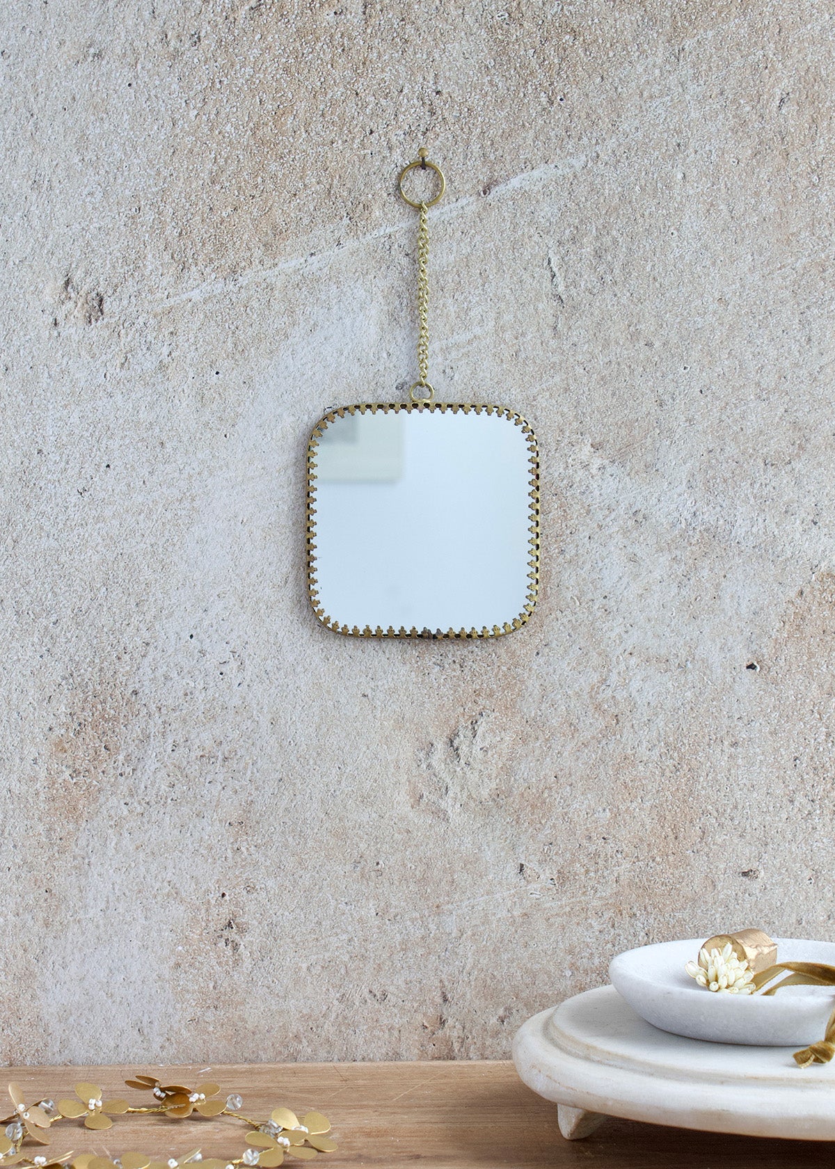 Small square mirror with rounded corners, brass edge and decorative chain for hanging, styled on a light stone wall with delicate accessories.
