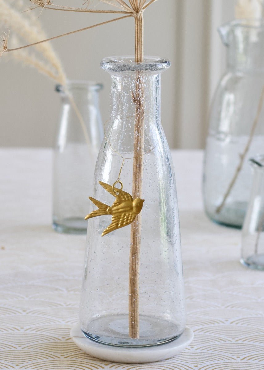 Golden swallow ornament hanging from a glass vase with dried flower stems in a serene table setting.