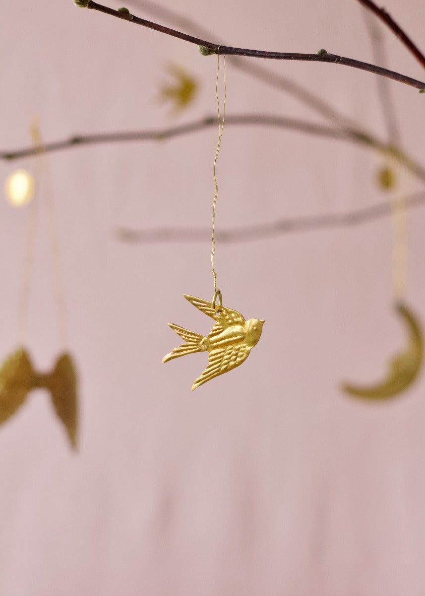Brass swallow ornament hanging from a spring branch with soft pink background and golden moon and wing shapes in the distance.