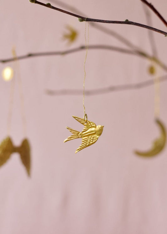 Brass swallow ornament hanging from a spring branch with soft pink background and golden moon and wing shapes in the distance.