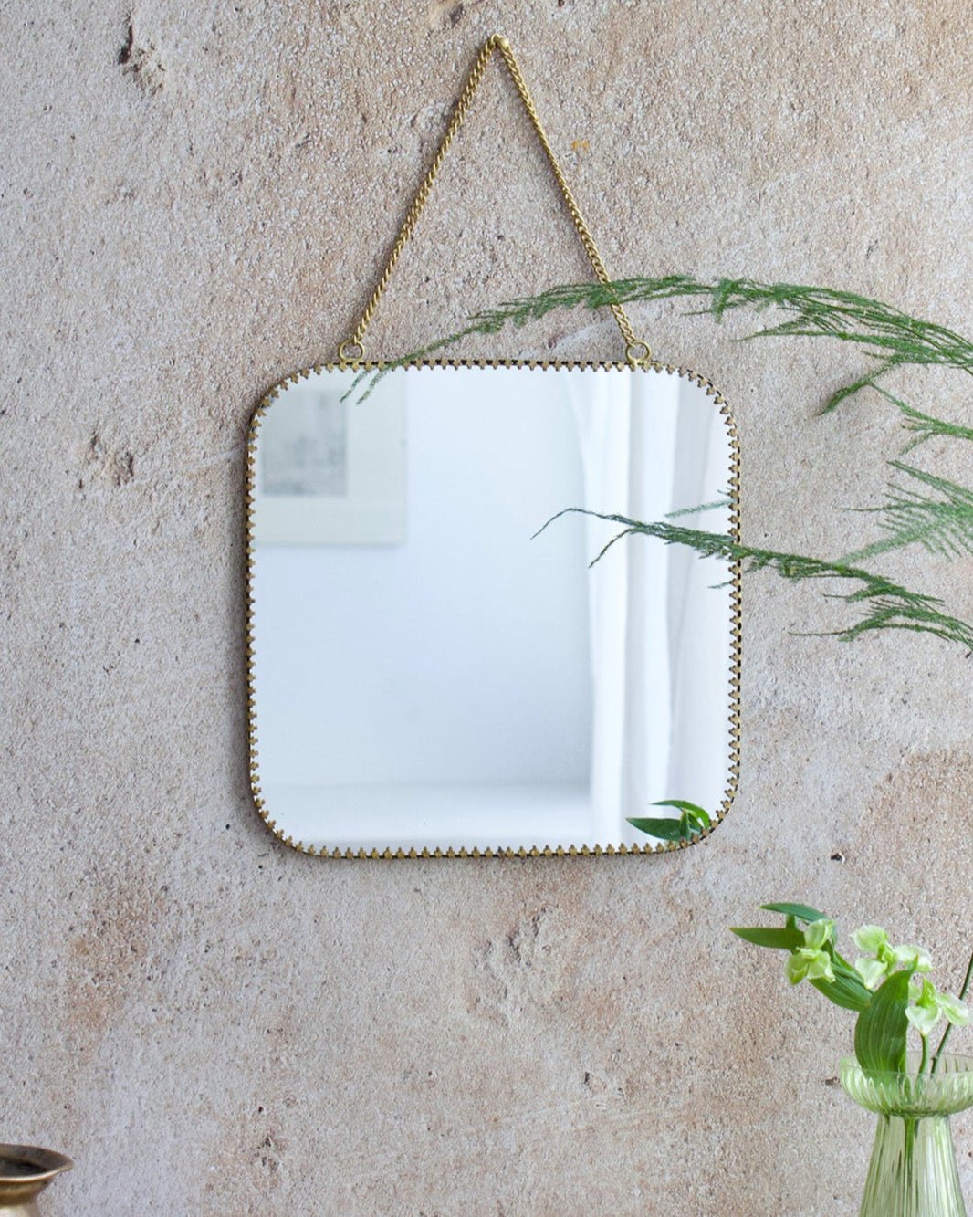 Square mirror with gold frame hanging on a textured wall with plants and decor items below.