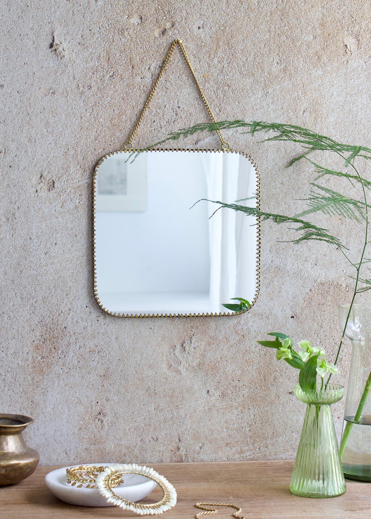 Square mirror with rounded corners and a brass scalloped edge, hanging from a brass chain on a rustic wall, styled with soft green flowers and vintage decor.