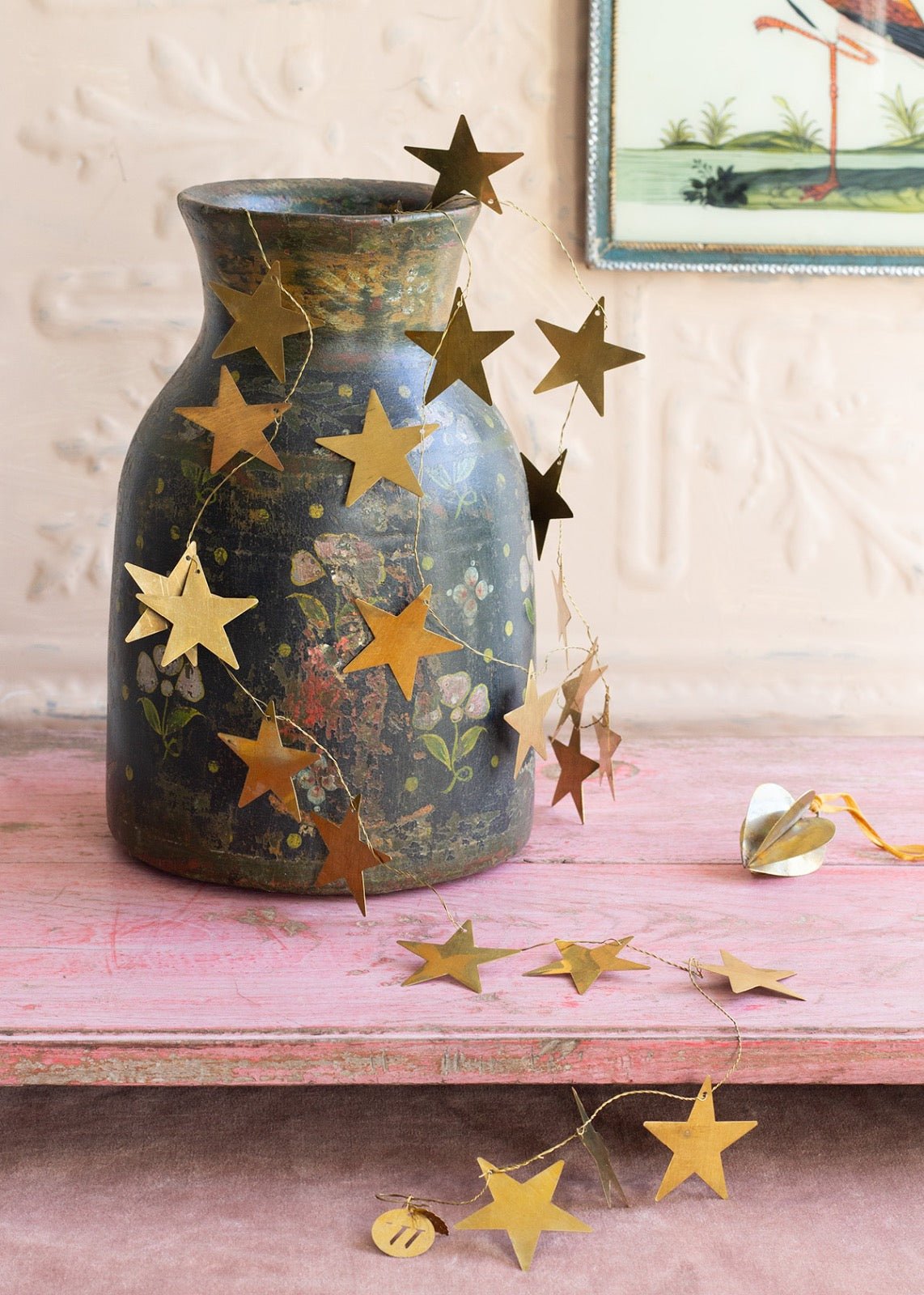 Golden brass star garland draped over a vintage painted vase with floral motifs, resting on a pink wooden surface with a soft pastel backdrop and a small heart ornament nearby.