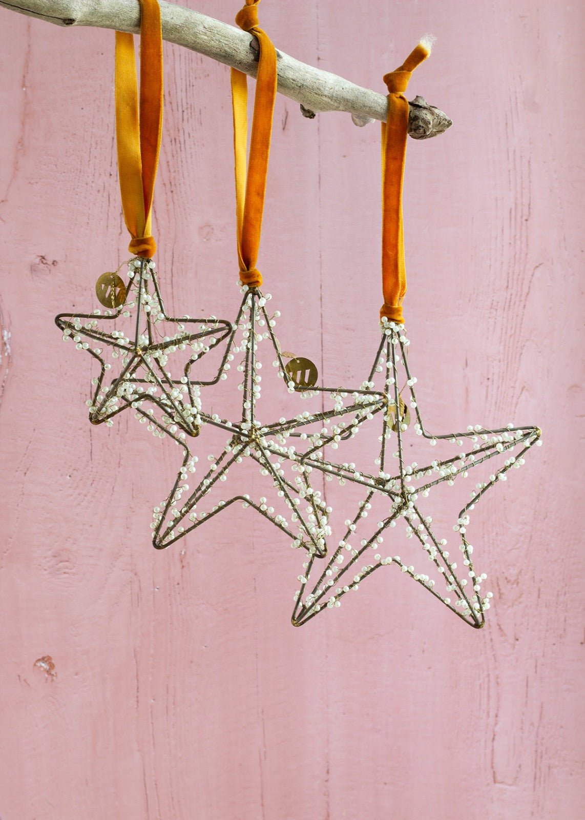 Three star-shaped wire ornaments decorated with tiny white beads, hanging from ochre velvet ribbons on a driftwood branch against a soft pink wooden background.