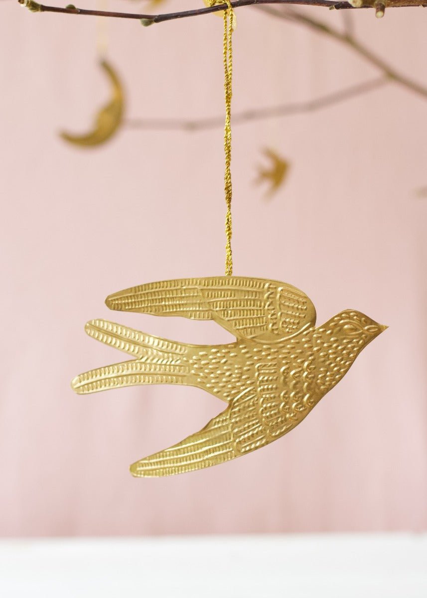 Brass swallow ornament hanging from a golden thread against a soft pink background, with blurred celestial shapes in the distance.