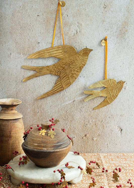 Two brass swallow wall ornaments, one large and one small, hanging from orange ribbons against a textured wall, with wooden pots and berry garland in the foreground.