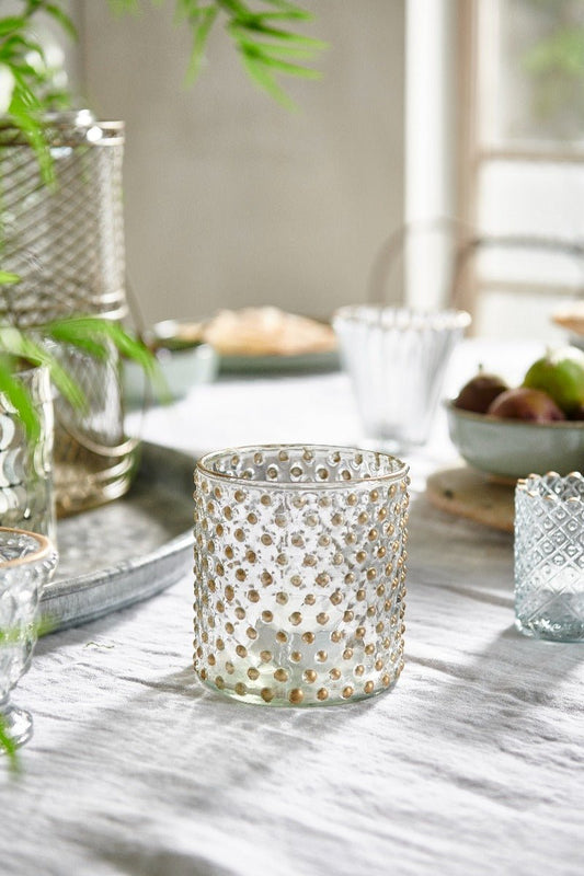 Glass tealight holder with gold hobnail texture on a softly lit dining table, surrounded by elegant glassware and a bowl of fresh fruit.