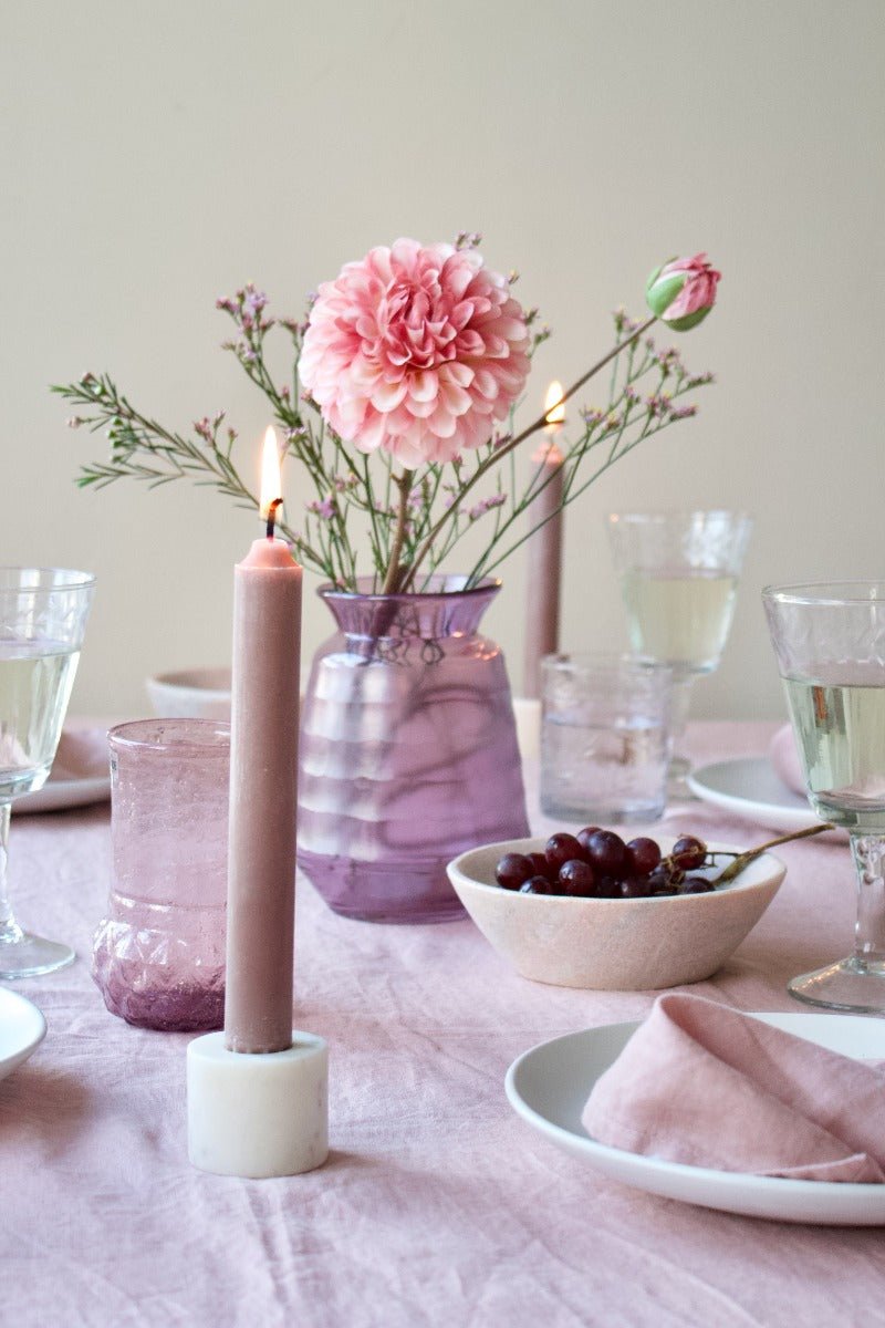 Lilac glass vase with pink flowers on a softly styled dining table, surrounded by wine glasses, a bowl of grapes, and lit pink candles.