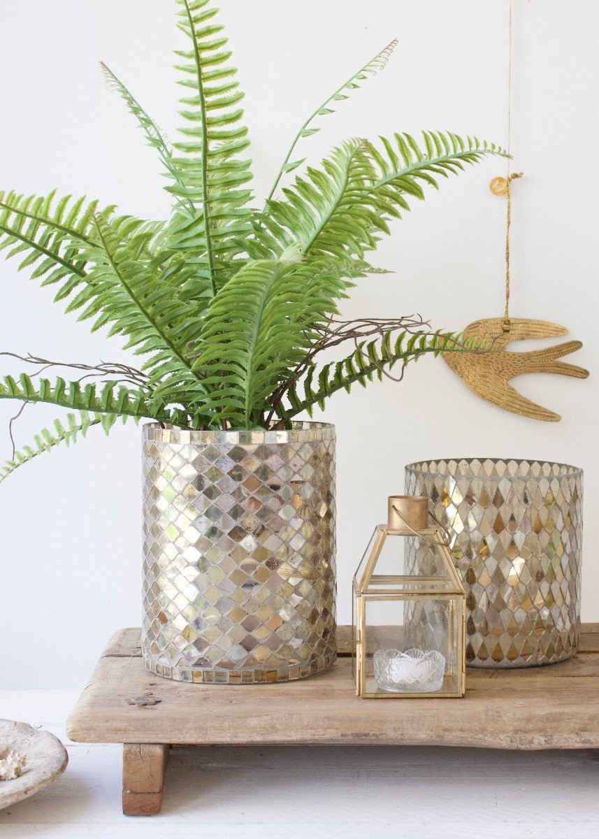 Handmade mosaic vase with golden and pearl-toned tiles, styled with a vibrant green fern and placed on a rustic wooden platform.