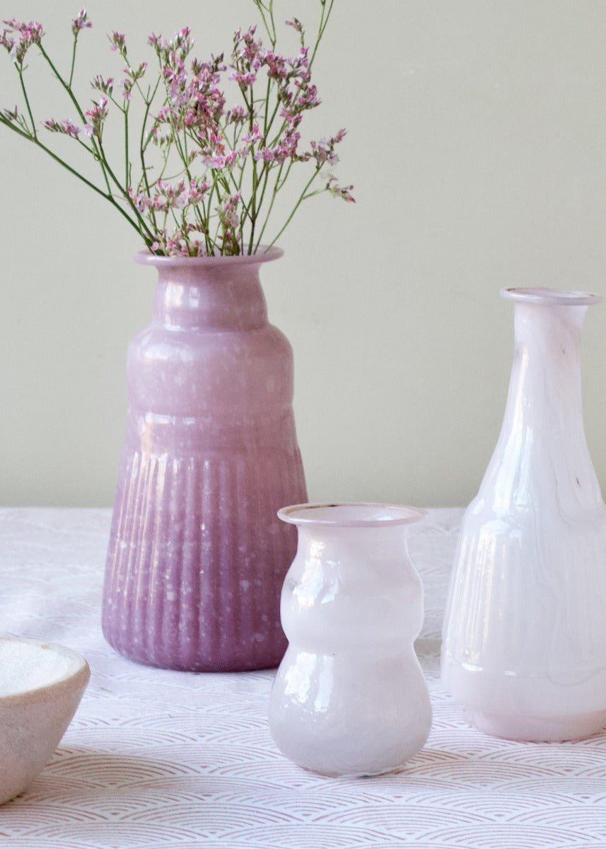 Tall lavender opaline glass vase with a flared rim and soft ribbed texture, styled with delicate pink wildflowers on a patterned tablecloth.