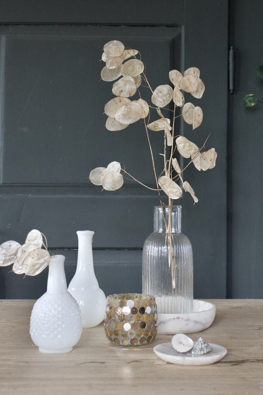 Elegant tabletop styling with two small white opaline vases, a ribbed glass vase with dried lunaria, a gold mosaic tealight holder, and white marble dishes with shells, set against a dark grey backdrop.