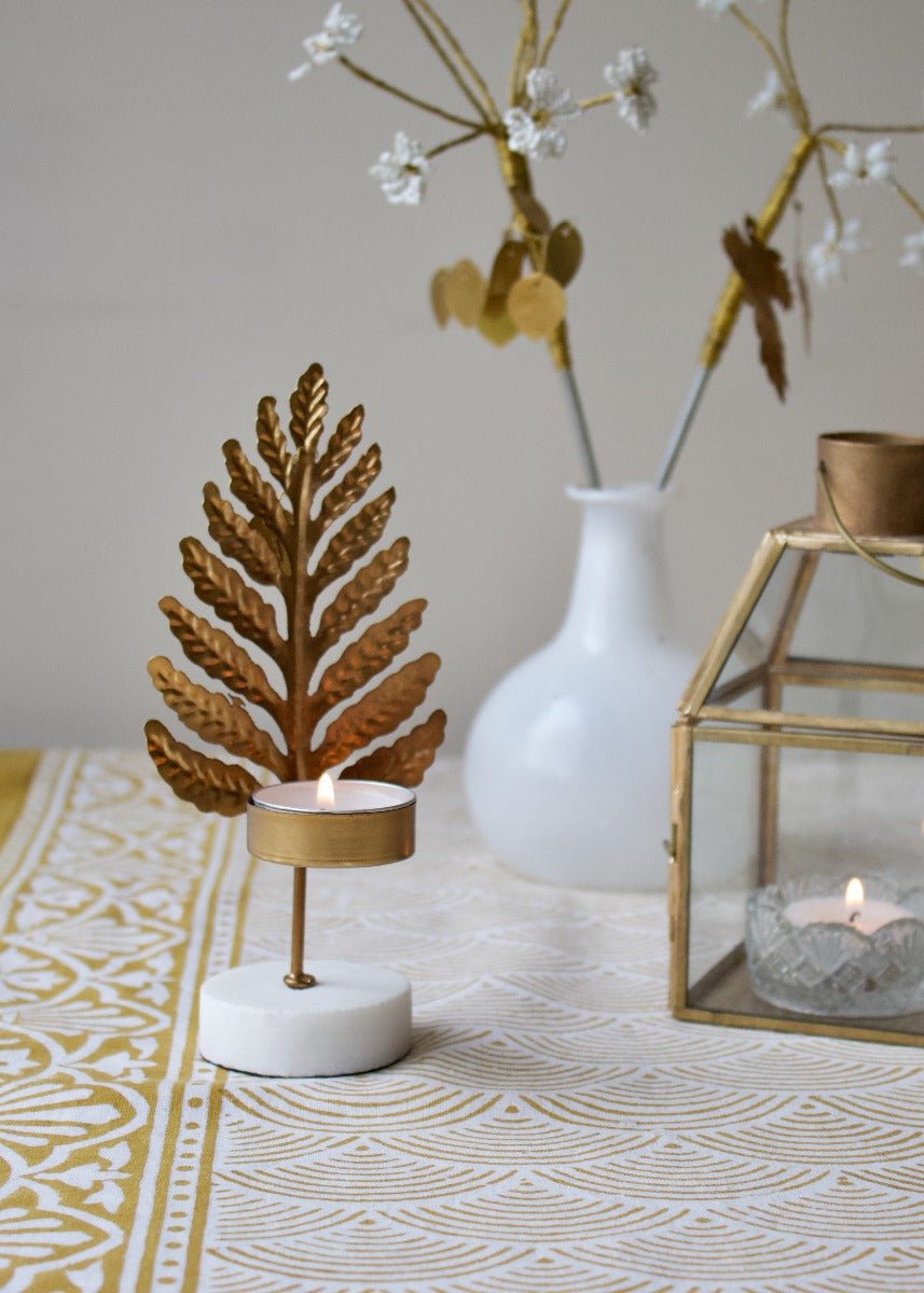 Gold-colored metal tealight holder in the shape of a stylized leaf, with a white round base, styled on a patterned yellow-and-white tablecloth.