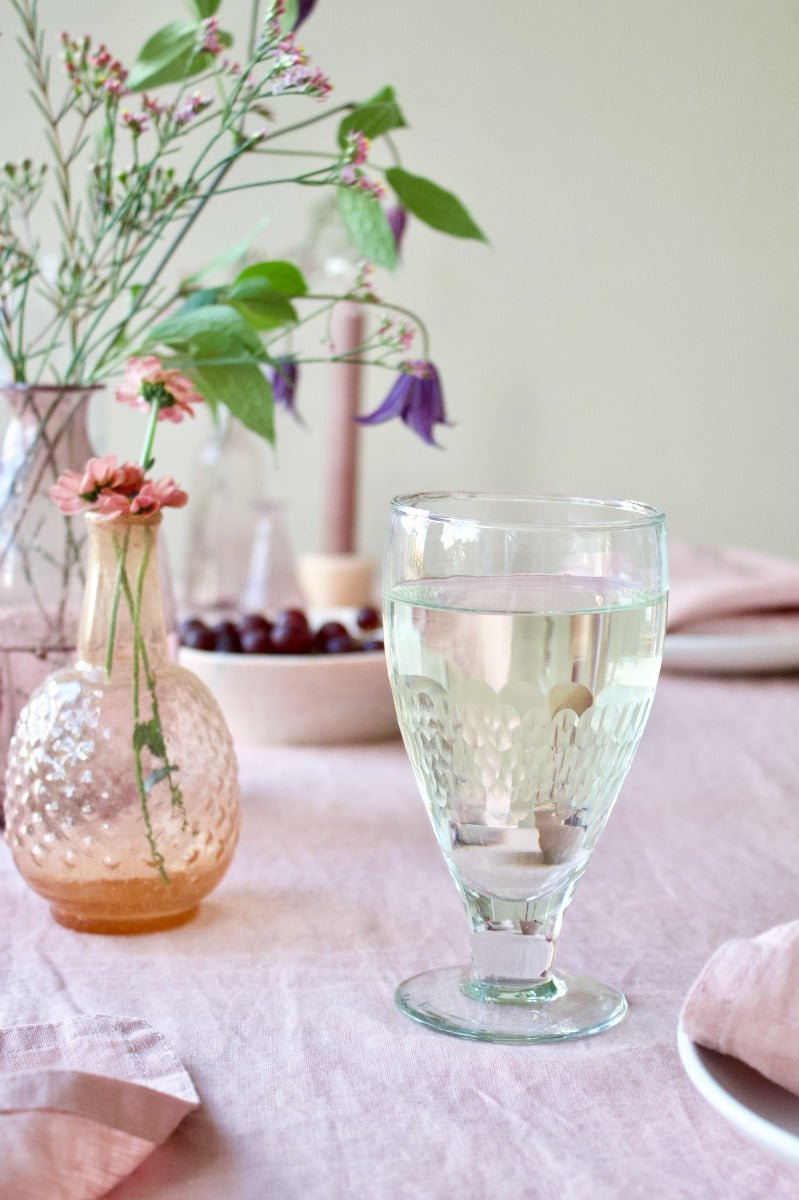 Close-up of a softly styled table setting with vintage glassware, including a pale amber vase holding small pink flowers and a footed drinking glass filled with water, on a blush pink linen tablecloth.