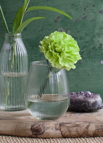 Small round ribbed glass vase with a wide base and narrow opening, styled with a single green carnation and placed on a wooden board next to a crystal.