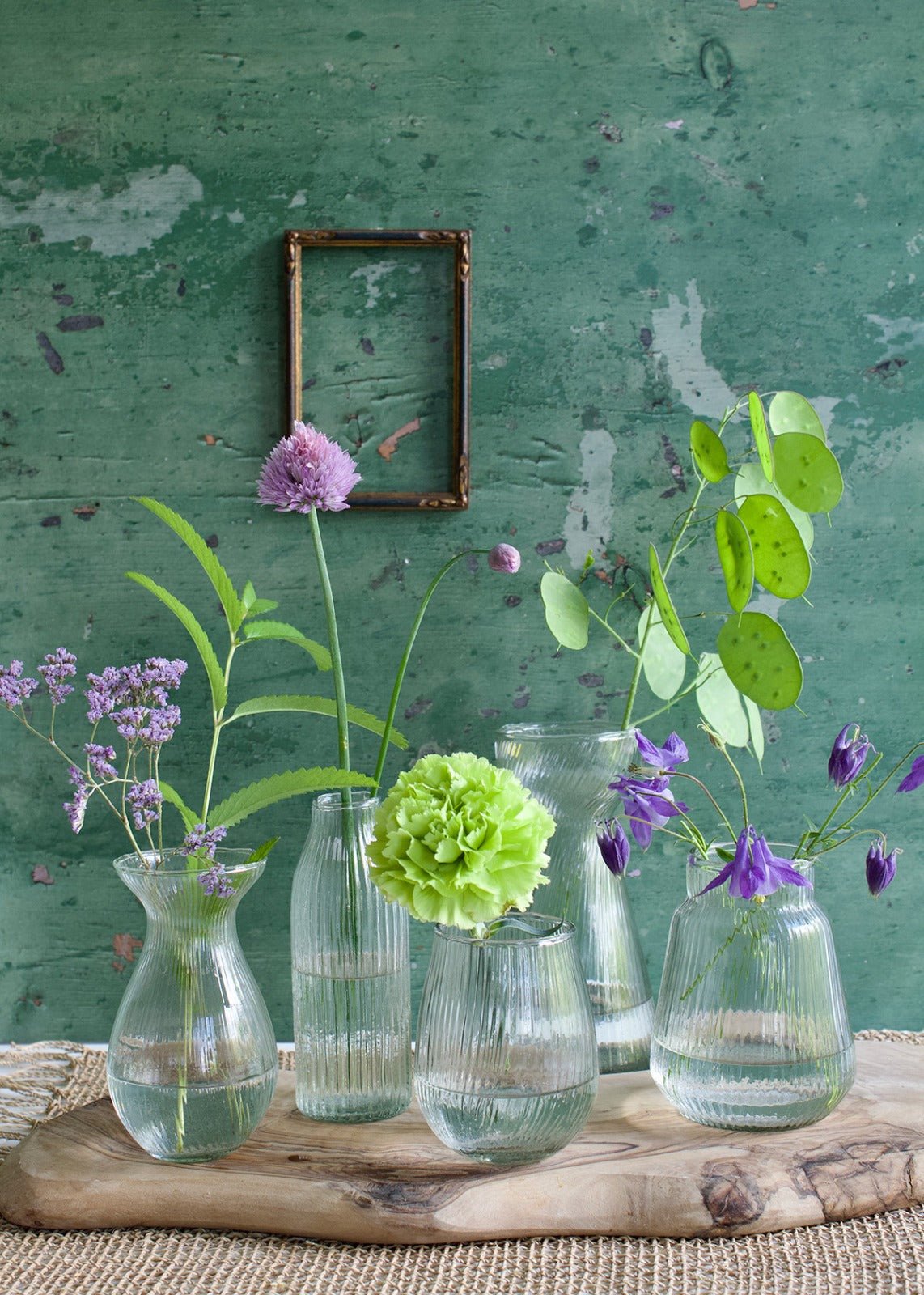 A set of five ribbed glass vases in various shapes and heights, each filled with water and seasonal wildflowers, styled on a rustic wooden board against a textured green wall.