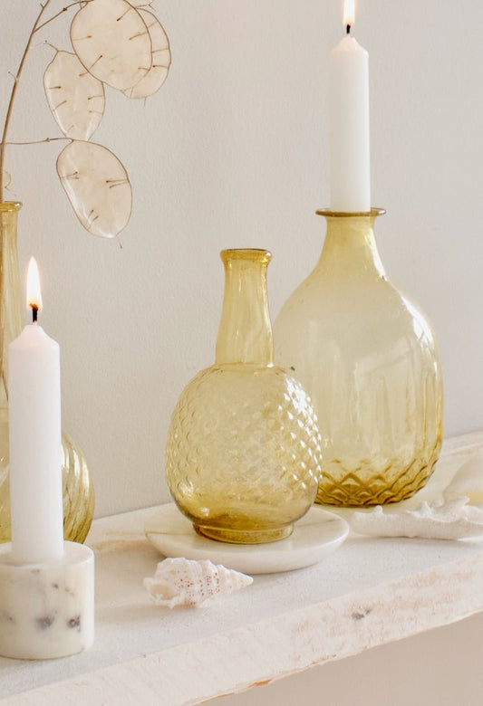 Amber glass vases with textured finishes, styled with lit white candles, dried lunaria branches and seashells on a rustic white shelf.