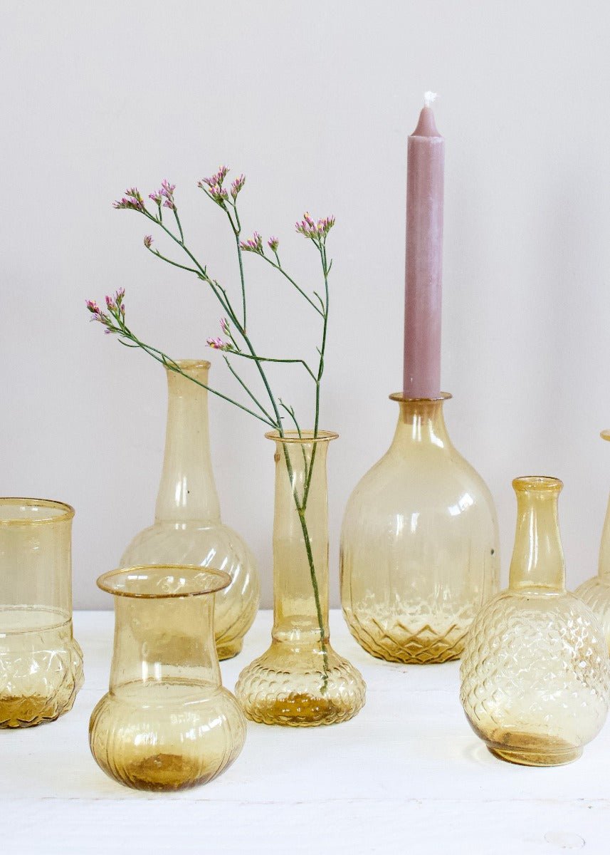 Close-up of amber glass vases in different textures and shapes, with a soft pink taper candle and delicate wildflowers, styled on a white wooden table.