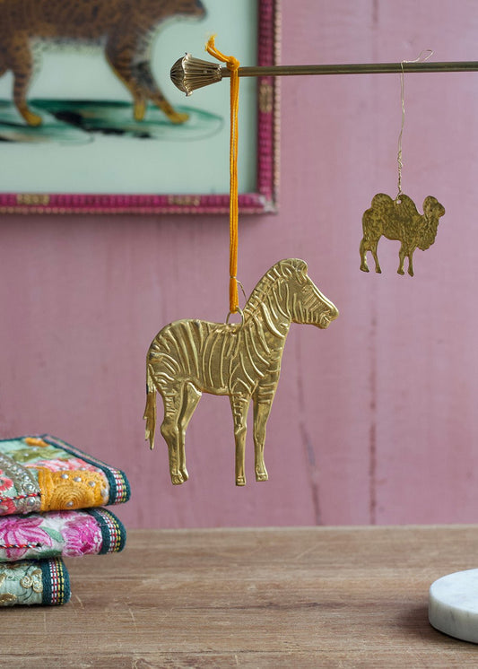 Brass zebra ornament hanging from a rod with velvet ribbon, accompanied by a small brass camel charm in the background, set against a soft pink wall.
