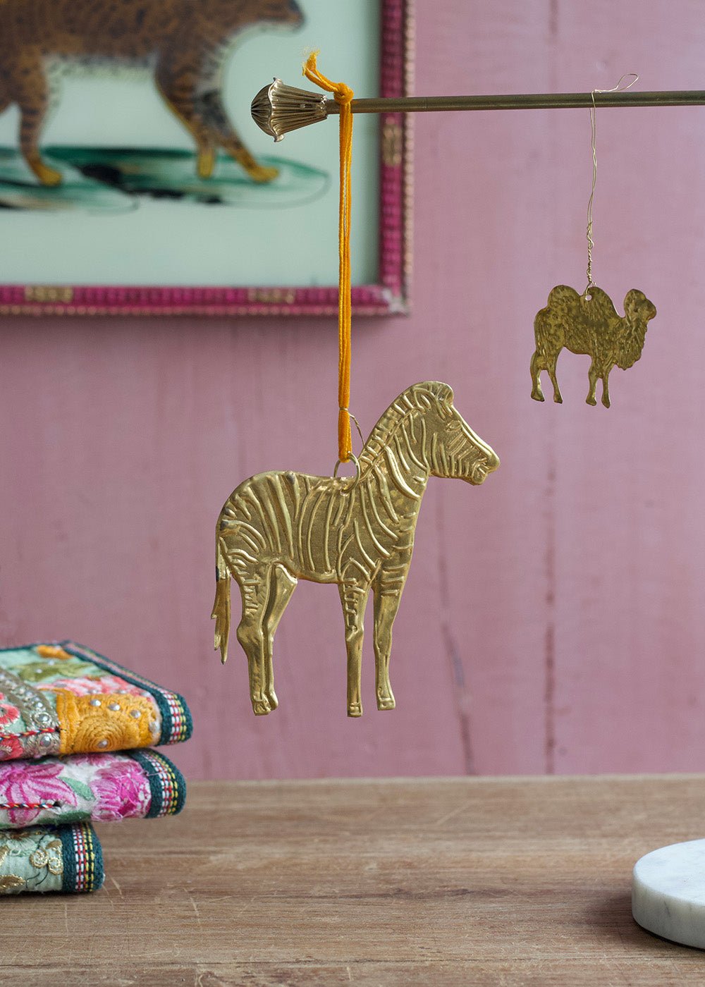 Brass zebra ornament hanging from a rod with velvet ribbon, accompanied by a small brass camel charm in the background, set against a soft pink wall.