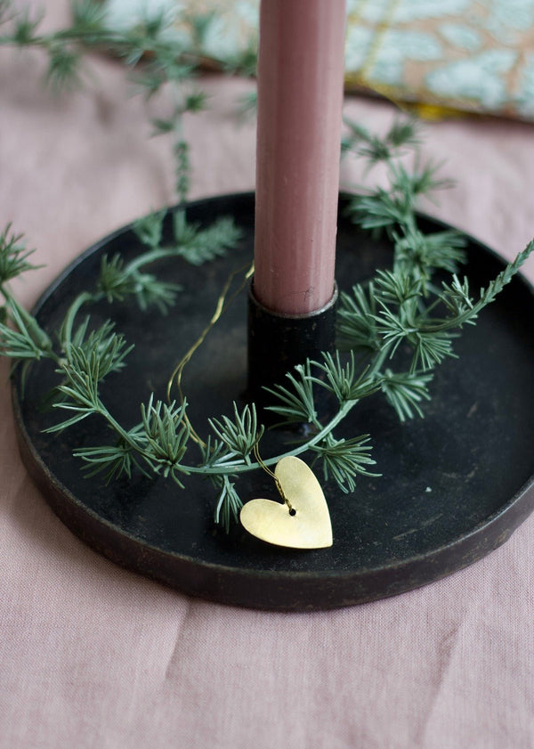 Small brass heart ornament hanging from a green garland around a pink candle on a dark metal base.