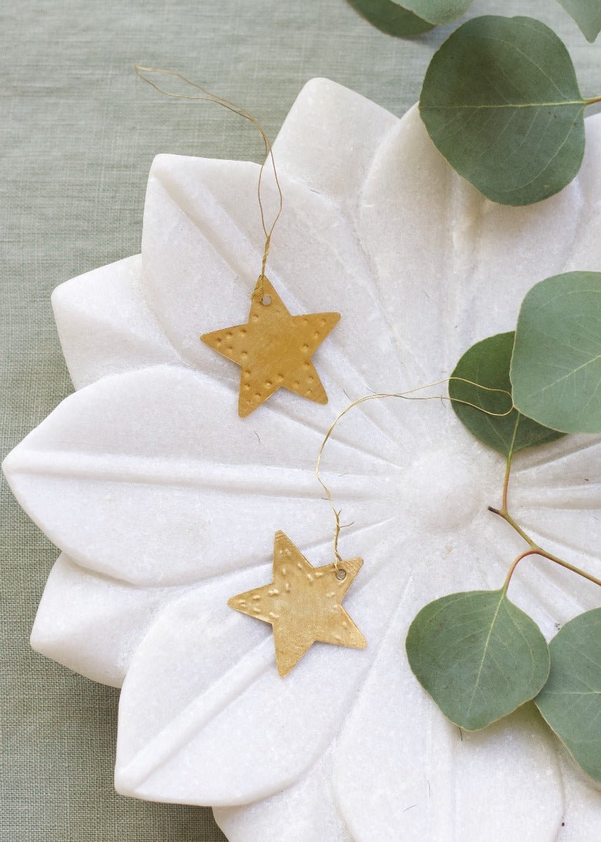 Two small gold star ornaments resting on a white carved flower decoration with eucalyptus leaves.

