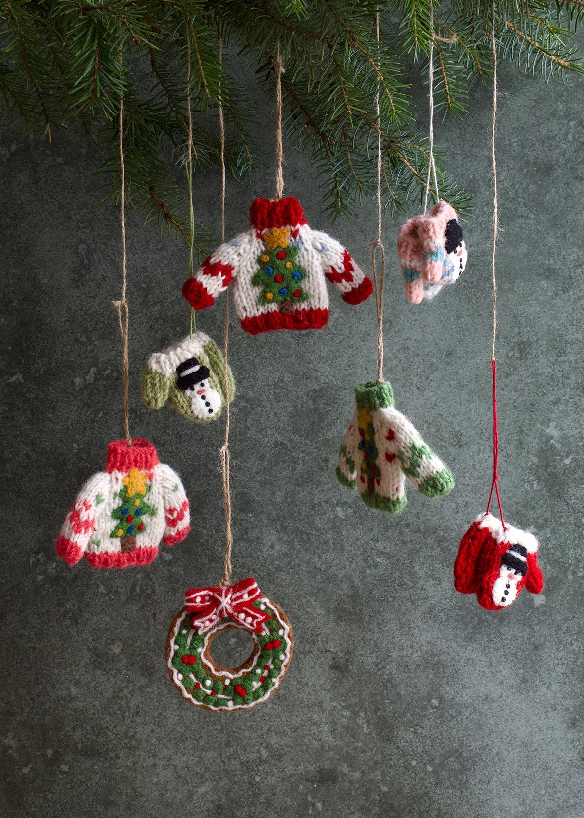 Knitted Christmas ornaments including sweaters, a wreath, and a snowman hanging against a dark background.