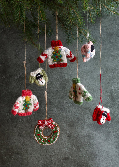 Knitted Christmas ornaments including sweaters, a wreath, and a snowman hanging against a dark background.