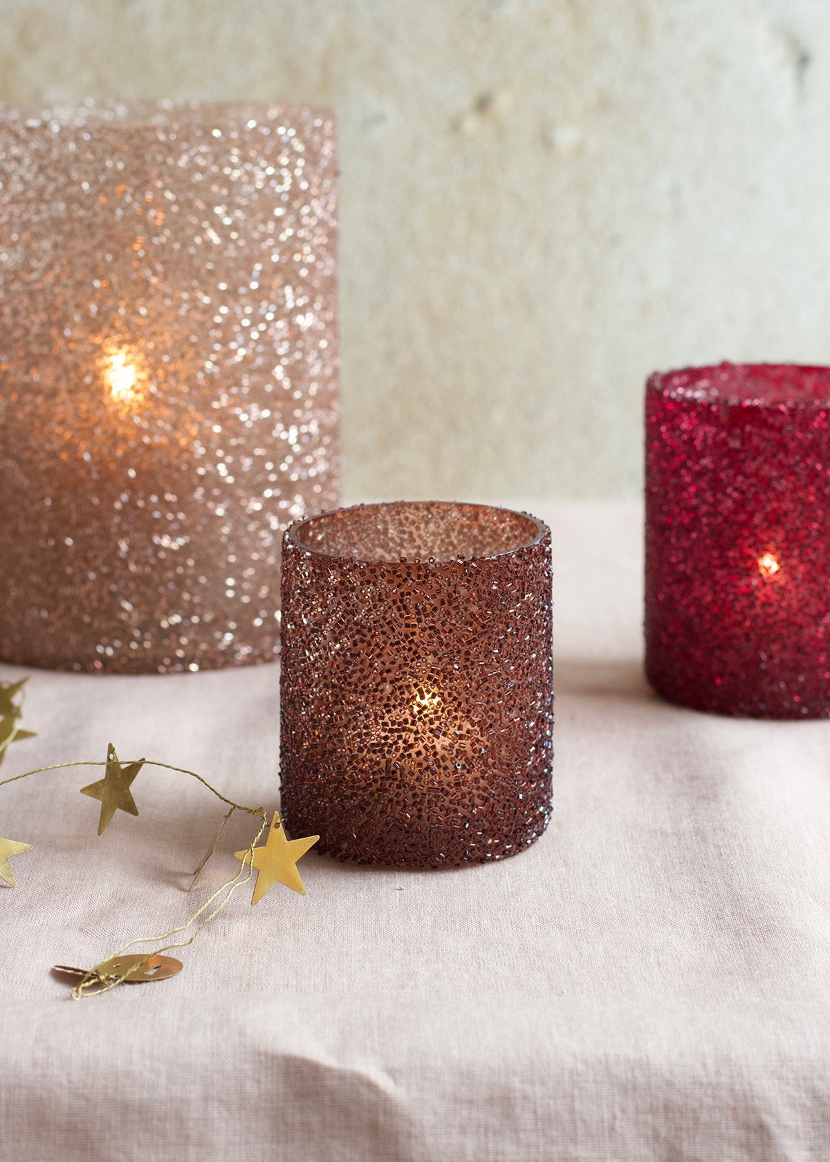 Three glittery candles in bronze, gold, and red on a textured surface with star-shaped decorations.