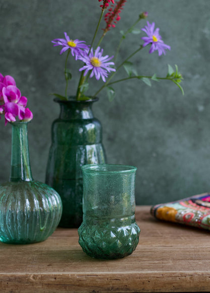 Green glass vase with textured surface on a wooden surface, with other vases and flowers in the background.