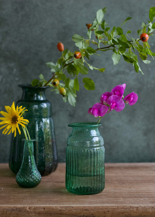 Three green glass vases with flowers on a wooden surface against a dark background