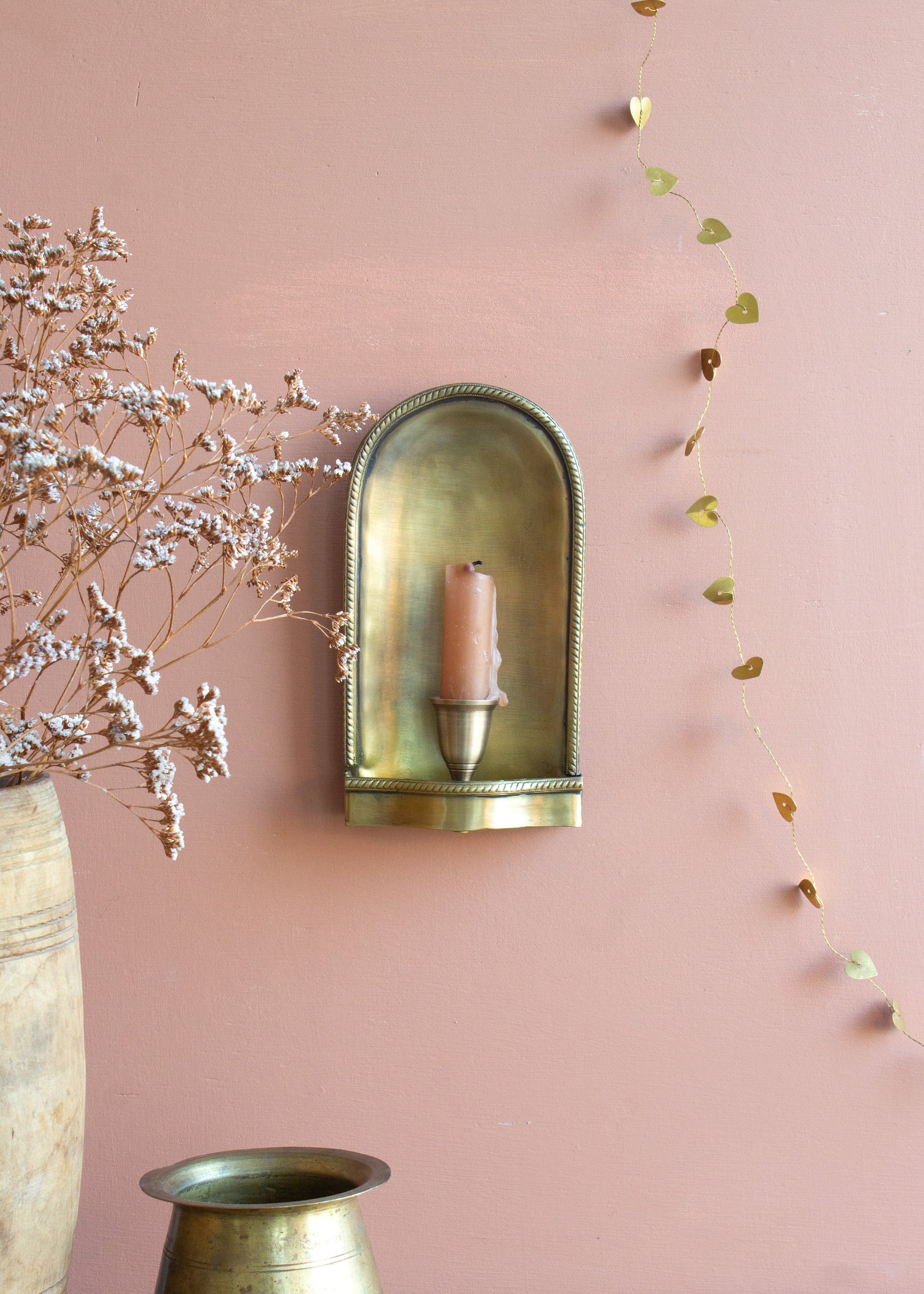 Brass wall sconce with a candle on a pink wall, decorated with dried flowers and greenery.