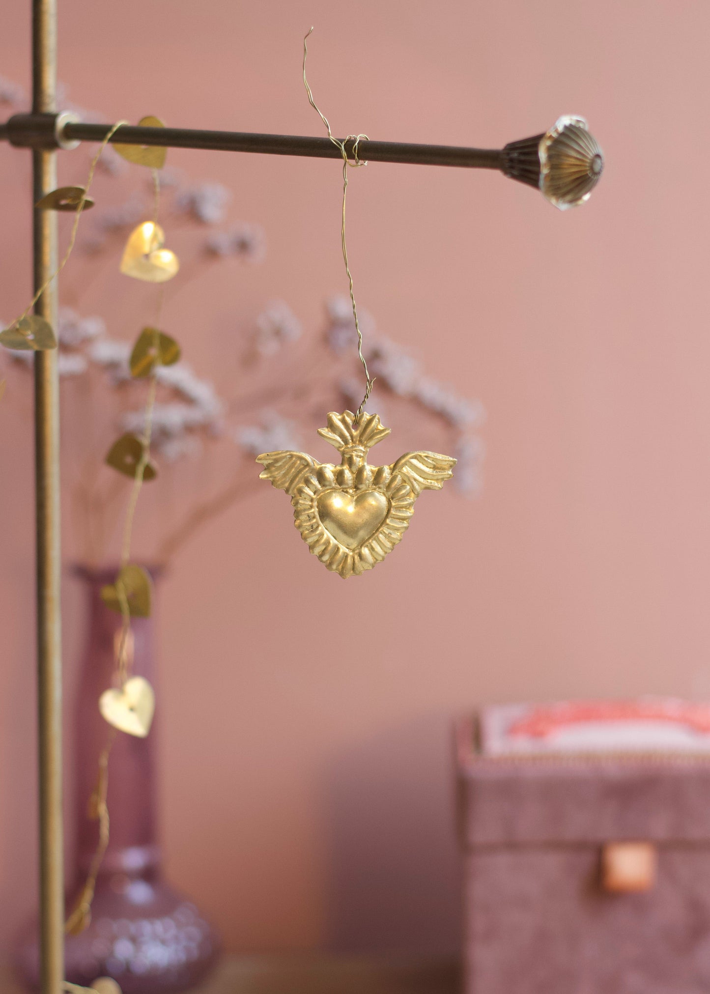 A gold heart-shaped ornament with wings hangs from a brass rod in a softly styled room with a pink wall, blurred decorative branches, and a purple vase.