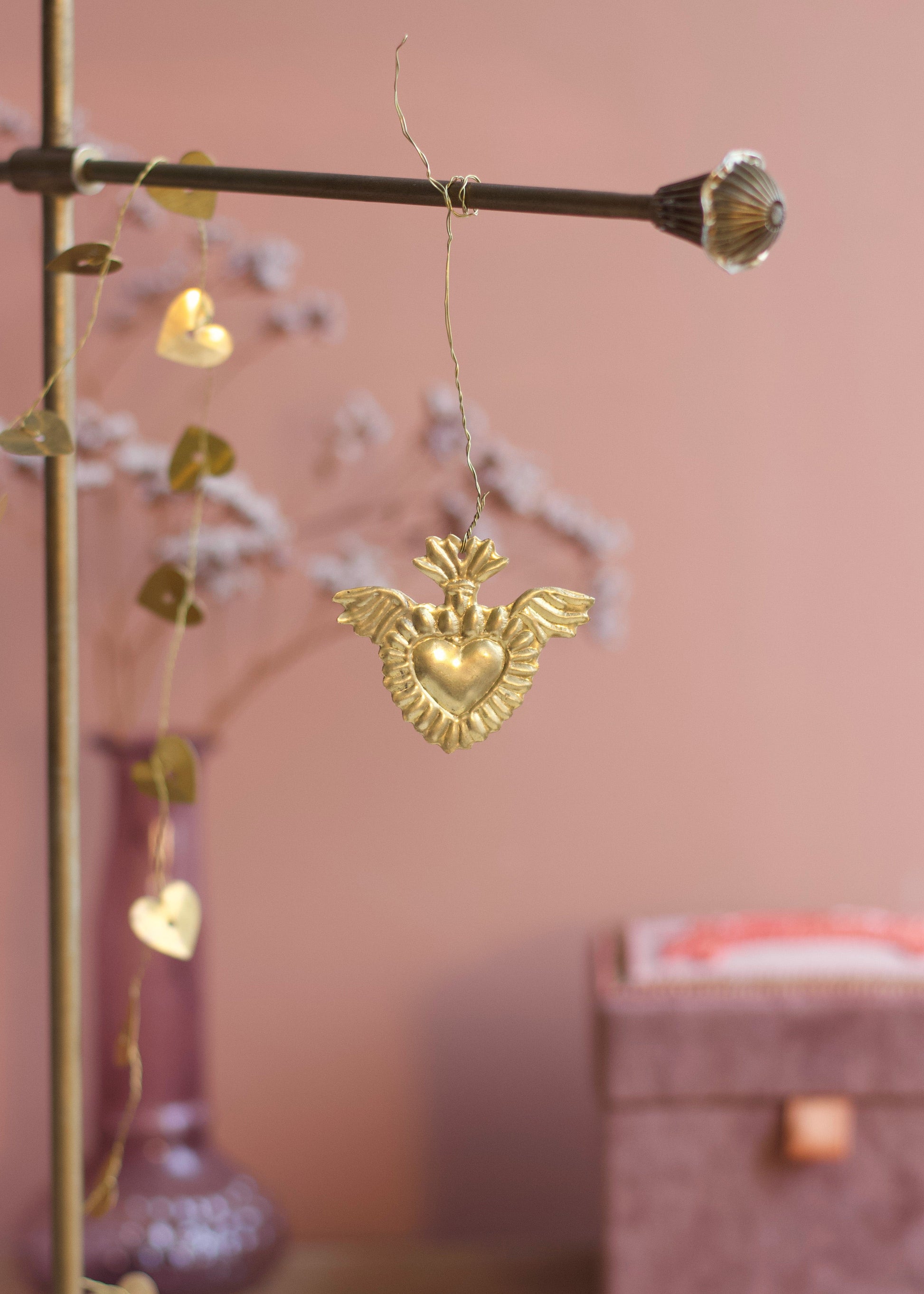 A gold heart-shaped ornament with wings hangs from a brass rod in a softly styled room with a pink wall, blurred decorative branches, and a purple vase.
