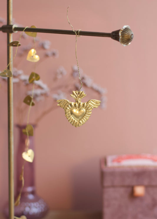 A gold heart-shaped ornament with wings hangs from a brass rod in a softly styled room with a pink wall, blurred decorative branches, and a purple vase.