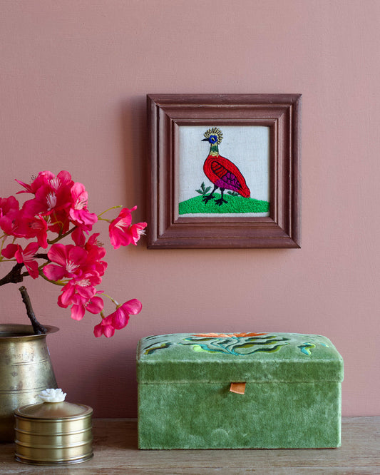 Framed embroidered artwork of a bird on a pink wall with a green box and pink flowers.